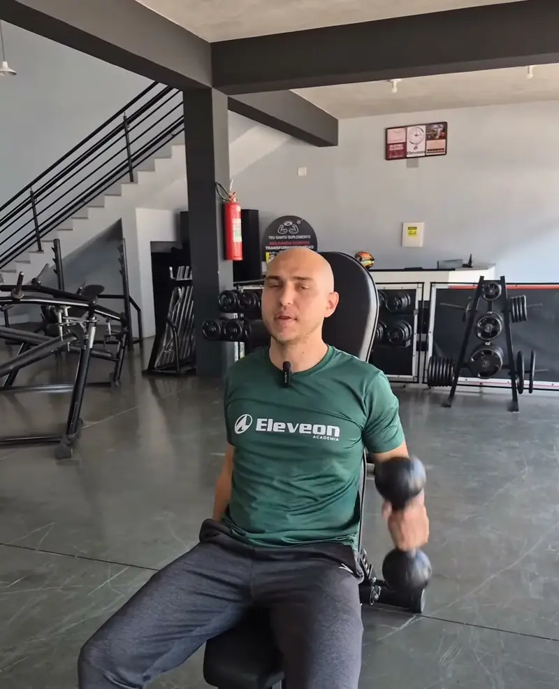 Professor da Eleveon sentado no banco, com halter na mão, na sala de musculação